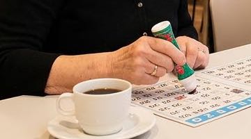 En äldre kvinna spelar bingo i en bingohall. Hon håller en grön bingopenna i handen och duttar numren på brickan. Det står en kopp kaffe intill.